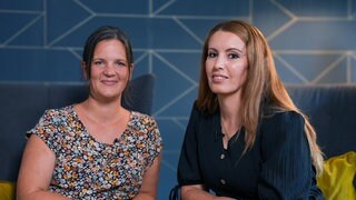 Karin und Peri leben mit ihren Familien zusammen im Hoffnungshaus. Sie sitzen vor einer dunkelblauen Wand mit glänzenden Mustern, nebeneinander auf zwei Sesseln. Karin hat ihre Haare in einem Pferdeschwanz und trägt eine geblümte Bluse. Peri trägt ihre langen, braunen Haare offen und ein schwarzes Sommerkleid. 