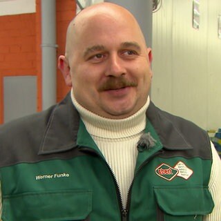 Werner Funke steht mit einer grünen Weste vor einer Lagerhalle. Er trägt einen hellbraunen Schnauzbart und eine Glatze.