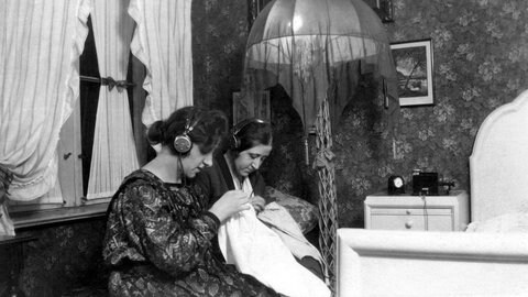 Zwei Frauen hören in den 1930ern mit Kopfhörer Radio und sticken: 1923 begann der reguläre Rundfunkbetrieb in Deutschland – maßgeblich vorangetrieben vom weitgereisten Ingenieur und Ministerialdirektor Hans Bredow