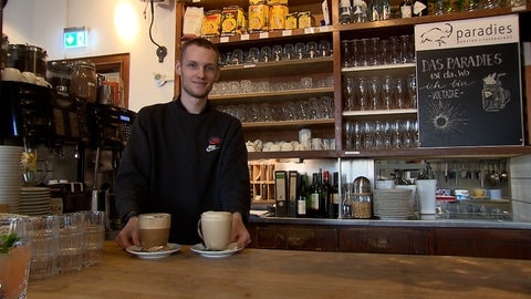 Ein junger Mann mit kurzen, blonden Haaren steht hinter einer Theke. Vor ihm stehen zwei Heißgetränke zum Servieren bereit.