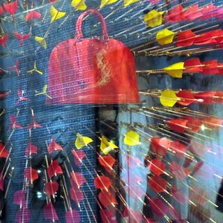 Eine rote Handtasche hängt in einem Schaufenster. Außen läuft eine Frau vorbei. Die Handtasche ist von kleinen Dartpfeilen in rot und gelb umgeben.