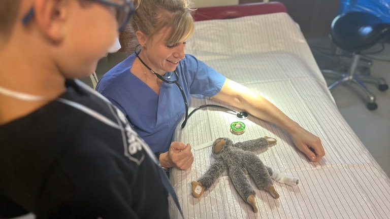 Plüschaffe liegt auf Behandlungsbett in einem Zimmer im Krankenhaus und wird von OP-Schwester versorgt.