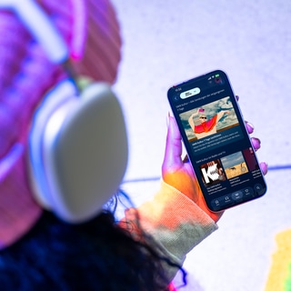 Eine Person mit Kopfhörern schaut auf ein Smartphone, auf dem das Angebot von SWR Kultur in ARD Sounds zu sehen ist