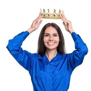 Eine Frau in Business-Kleidung setzt sich selbst lächelnd eine Krone auf: Studien zeigen, dass zumindest weibliche narzisstische CEOs die Unternehmensleistung steigern. 