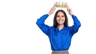 Eine Frau in Business-Kleidung setzt sich selbst lächelnd eine Krone auf: Studien zeigen, dass zumindest weibliche narzisstische CEOs die Unternehmensleistung steigern. 