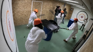 4 Frauen halten Baseballschläger in der Hand und schlagen auf einen Fernseher ein. Sie tragen weiße Overalls und einen orangenen Schutzhelm.