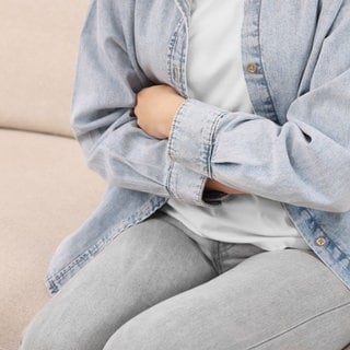 Eine Frau sitzt auf einem Sofa und hält sich den Magen. Nach zahlreichen Magen-Darm-Erkrankungen nach einem Landfrauenfest steht das Norovirus als Ursache fest.