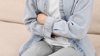 Eine Frau sitzt auf einem Sofa und hält sich den Magen. Nach zahlreichen Magen-Darm-Erkrankungen nach einem Landfrauenfest steht das Norovirus als Ursache fest.