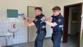Zwei Polizisten gehen mit gezogenen Übungswaffen und Taschenlampen durch einen Raum.