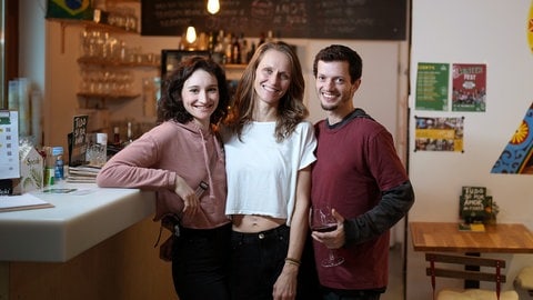 Clara, Imogen und Jadu leben polyamor. Sie stehen in ihrem Restaurant und lächeln in die Kamera.