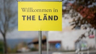 An einem Ortsschild hängt ein Zusatzschild mit der Aufschrift "Willkommen in THE LÄND2.