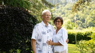 Altes Paar im Garten, das sich in den Armen hält. Beide tragen ein weißes Oberteil.