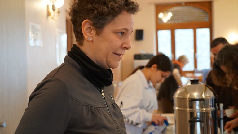 Frau mit grauen Locken im Profil, im Hintergrund Menschen.