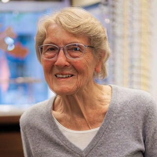 Älter Dame mit blonden kurzen Haaren lächelt in die Kamera. Im Hintergrund erkennt man einen Brillenladen. 