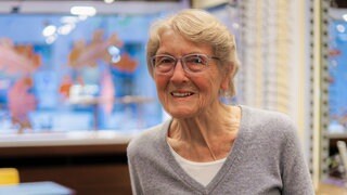 Älter Dame mit blonden kurzen Haaren lächelt in die Kamera. Im Hintergrund erkennt man einen Brillenladen. 
