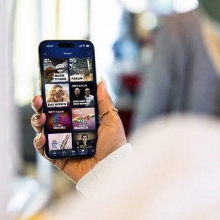 Eine Person mit Kopfhörern schaut auf ein Smartphone, auf dem das Angebot von SWR Kultur in ARD Sounds zu sehen ist