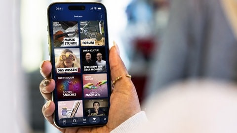 Eine Person mit Kopfhörern schaut auf ein Smartphone, auf dem das Angebot von SWR Kultur in ARD Sounds zu sehen ist