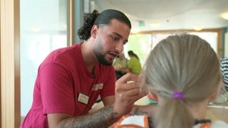 Der Auszubildende Kaan Alkan reicht einer älteren Dame mit einem Löffel das Mittagessen an.