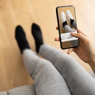 Eine Person probiert zu Hause auf dem Sofa mithilfe der App eines Versandhändlers virtuell neue Schuhe an: Wenn Kunden beim Online-Shopping mithilfe von KI passgenaue Mode auswählen können, soll das helfen, Retouren zu vermeiden.