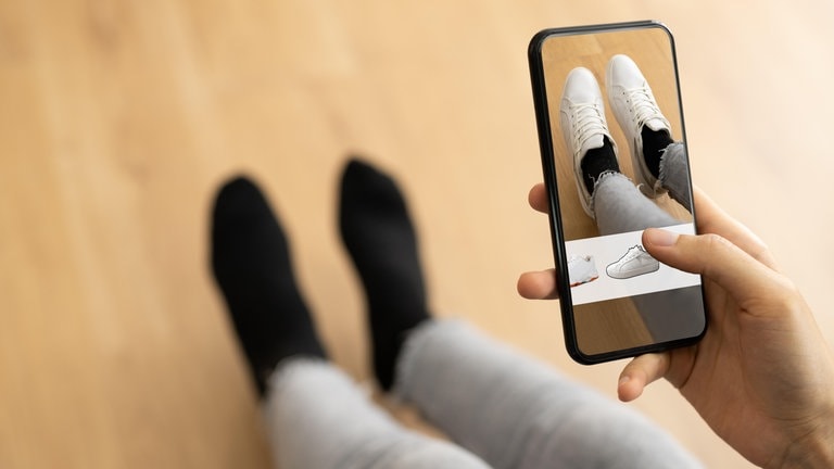 Eine Person probiert zu Hause auf dem Sofa mithilfe der App eines Versandhändlers virtuell neue Schuhe an: Wenn Kunden beim Online-Shopping mithilfe von KI passgenaue Mode auswählen können, soll das helfen, Retouren zu vermeiden.