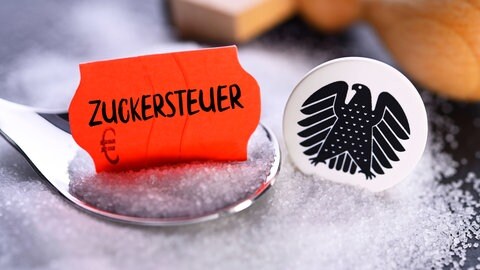 Symbolbild: Ein Löffel voll Zucker mit einem Schild, auf dem "Zuckersteuer" steht, daneben ein Bundesadler und ein Stempel