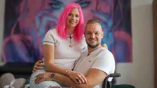 Mathias ist querschnittsgelähmt und sitzt seit 4 Jahren im Rollstuhl. Seine Partnerin Lisa hat pinke Haare und sitzt auf seinem Schoß. Sie umarmen sich und lachen in die Kamera. 