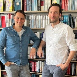 Die Journalisten Roland Peters und Hannes Vogel stehen gemeinsam vor einem Bücherregal.