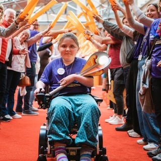 Die junge Fackelträgerin fährt im Rollstuhl bei der Auftaktversanstaltung des Kinder-Lebens-Laufs durch ein Spalier an Unterstützenden.