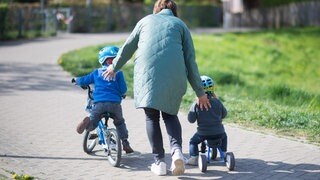 Symbolbild | Eine Mutter schiebt ihre kleinen Kinder an, die mit Fahrrad und Tretroller unterwegs sind. 