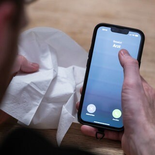 Ein Mann hält ein Taschentuch in einer Hand und ein Telefon mit dem Schriftzug "Arzt" in der Anderen. 