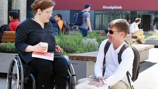 Eine Studentin im Rollstuhl und ein blinder Student mit Blindenhund unterhalten sich auf dem Campus einer Uni: Menschen mit Behinderungen sollen gleichberechtigten Zugang zur Hochschulbildung haben. In der Praxis sind Seminarräume mit Rollstühlen aber oft schlecht erreichbar und Studienordnungen eng getaktet. 