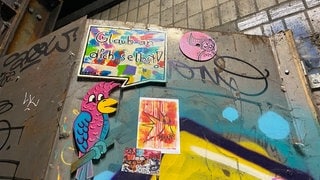 Auf einem stählernen Gerüst sind Aufkleber, eine bunte Fliese mit der Aufschrift "Glaube an dich selbst", ein Papagei und eine pinkfarbene Platte angebracht.