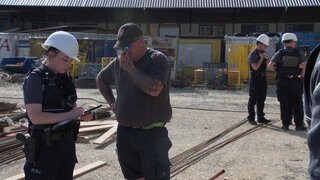 Eine Zollbeamtin notiert sich etwas in einen Block, während der Baustellenleiter daneben steht und sich im Gesicht kratzt. Im Hintergrund stehen unscharf weitere Zollbeamte.