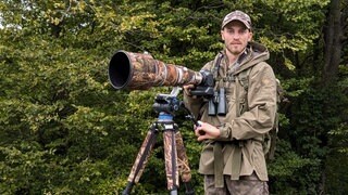 Ein junger Mann in Tarnkleidung steht vor einer Kamera in Tarnfarben. Er ist Tierfotograf.