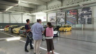 Sonja Faber-Schrecklein steht mit zwei Mitarbeitern des Technikmuseums Sinsheim in einer Autohalle. Links von ihnen stehen Oldtimer.