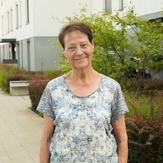 Eine ältere Frau mit braunen Haaren lächelt in die Kamera. Hinter ihr ist ein weißer Wohnkomplex mit vielen Bäumen rundherum zu sehen.