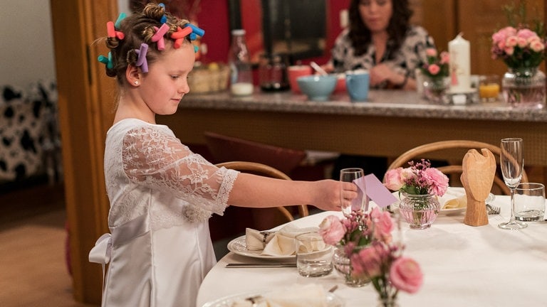 Carlotta in ihrem Kommunionkleid und Papilotten in den Haaren am festlich gedeckten Tisch