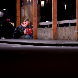 Eine obdachlose Frau sitzt in Decken eingehüllt an einem Bahnhof.