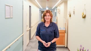Heike ist Palliativkrankenschwester und steht in der Mitte eines hellen Flurs des Hospizes, neben ihr steht ein Strauß mit lila Tulpen. 