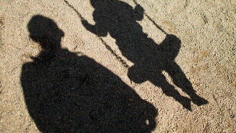 Der Schatten von einem Mann und einem schaukelnden Kind sind im Sand auf einem Spielplatz zu sehen. Ein Tagesvater im Rems-Murr-Kreis soll Kinder schwer sexuell missbraucht haben.