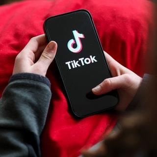 Ein Mädchen hält ein Smartphone, auf dem das TikTok-Logo zu sehen ist: Auf Social Media stoßen Kinder und Jugendliche schnell auf problematischen Content: Extremismus, Gewalt, Essstörungs-Communitys. Was braucht es für einen digitalen Jugendschutz?
