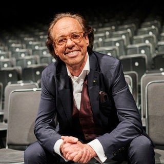 Kabarettist Christoph Sonntag sitzt in einem Raum voller Stühle in der ersten Reihe.