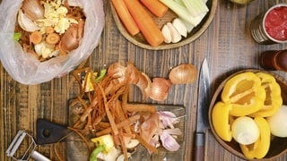 Frisches Gemüse und Küchenabfälle liegen auf einem Holztisch. Neben Schalen und einem Messer gibt es Karotten, Sellerie, Paprika und Knoblauch. Einige Artikel befinden sich in Schalen und einem Plastikmüllbeutel. Biomüll wie Zwiebelschalen und Kaffeesatz eignen sich zum natürlichen Düngen.
