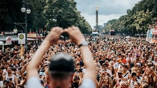 Ein Mann steht vor einer Menschenmenge und macht mit seinen Händen ein Herzzeichen, im Hintergrund ist die Berliner Siegessäule zu sehen