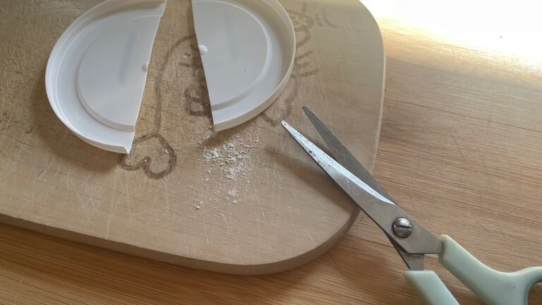 Den Deckel mit der Schere mittig gerade durchschneiden. Die beiden Hälften werden nun mit Heißkleber von außen so an den Ring geklebt, dass sie genau gegenüber liegen. 