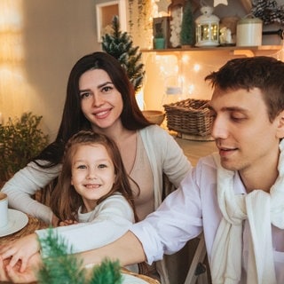 Eine junge Familie sitzt gemütlich und zufriedend lächelnd an einem Tisch inmitten von Weihnachtsdekoration