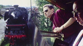Blick auf eine Dampflok und einem Mann mit Sonnenbrille, der während der Fahrt aus einem Bahnabteil schaut.