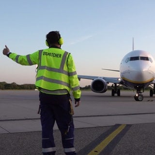 Simon Fauth steht mit einer neongelben Weste mit der Aufrschrift "Dispatcher" vor einem Flugzeug und zeigt mit dem linken Daumen nach oben.
