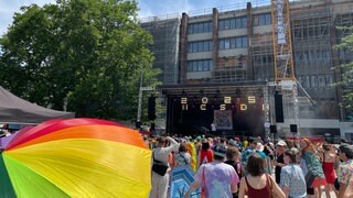 Im Vordergrund prankt ein Regenbpgen-Regenschirm. Der Christopher Street Day (CSD) starte im Freiburg mit einer Kundgebung auf dem Platz der Alten Synagoge. Im Hintergrund spricht Oberbrügermeister Martin Horn auf der Bühne.