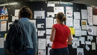 Zwei Studentinnen betrachten am die Wohnungsanzeigen am Schwarzen Brett.